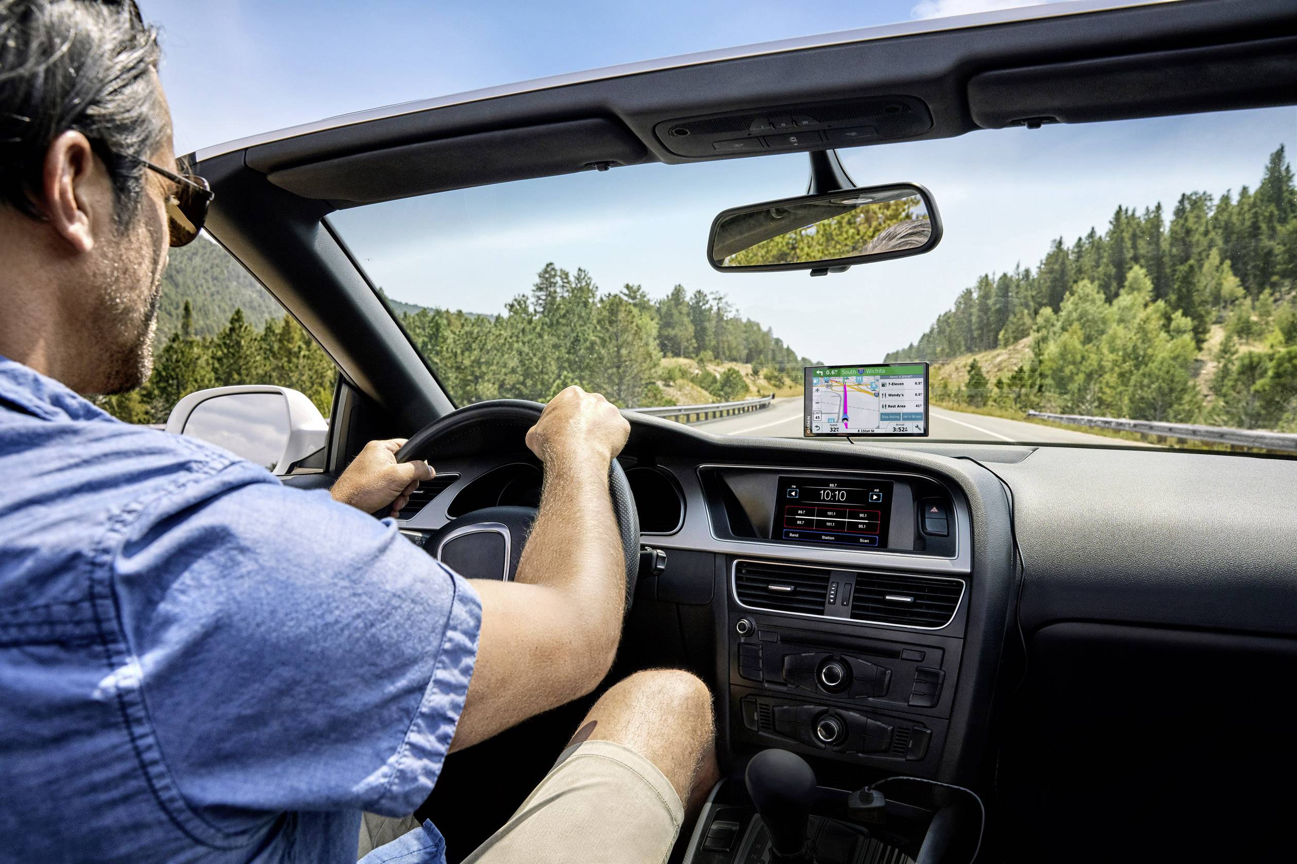 A man is driving a car on a country road while looking at the GPS navigation device. Wooded hills are visible in the background.