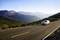 A silver car drives along a winding road through a mountainous landscape with wooded hills and a blue sky in the background.