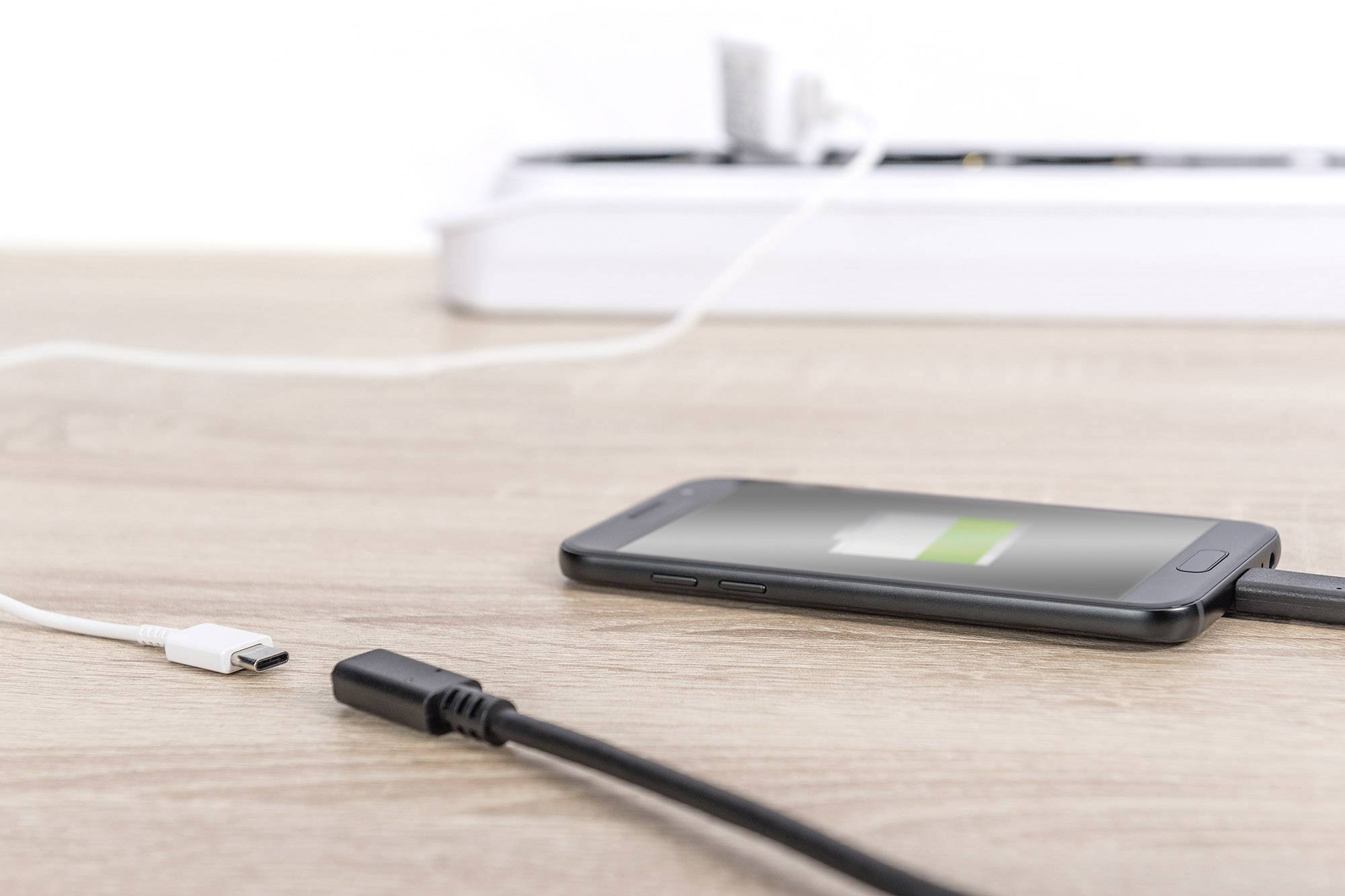 A mobile phone is being charged on a wooden table. In the background, a power strip with plugged-in cables can be seen.