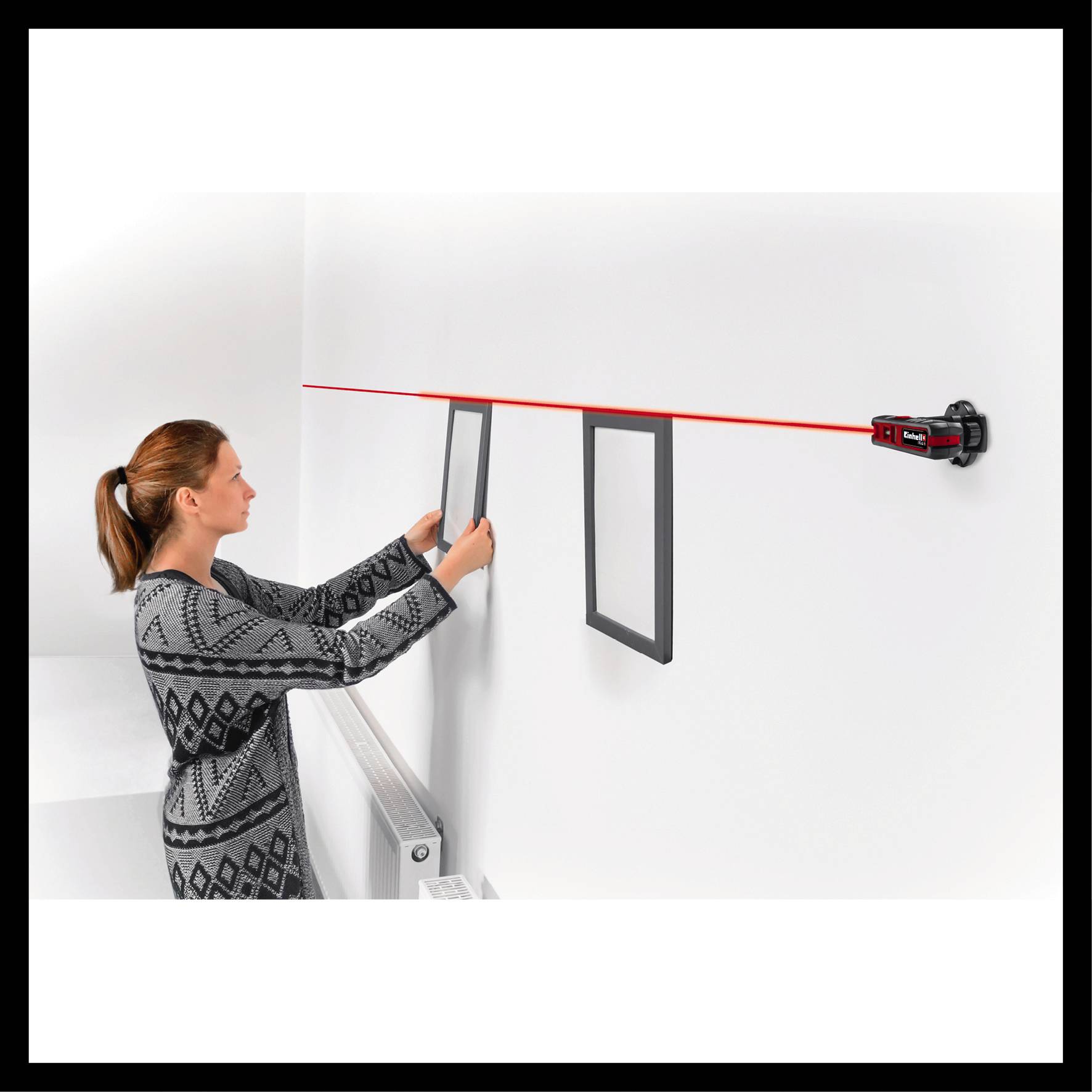 A woman is hanging two pictures on a white wall using a laser level.