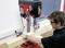 A man is using a pillar drill to work on a large piece of wood in a workshop. Safety precautions are visible.