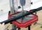 A metal piece is secured on a red and black drill stand. A protective housing surrounds the drill, which is positioned above the workpiece.