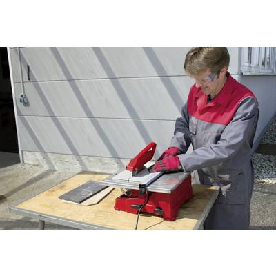 A person is cutting tiles with a red tile cutter on an outdoor table. They are wearing safety glasses and gloves.