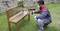 A person is painting a wooden bench in the garden using a spray gun. They are wearing protective clothing and a respirator mask.