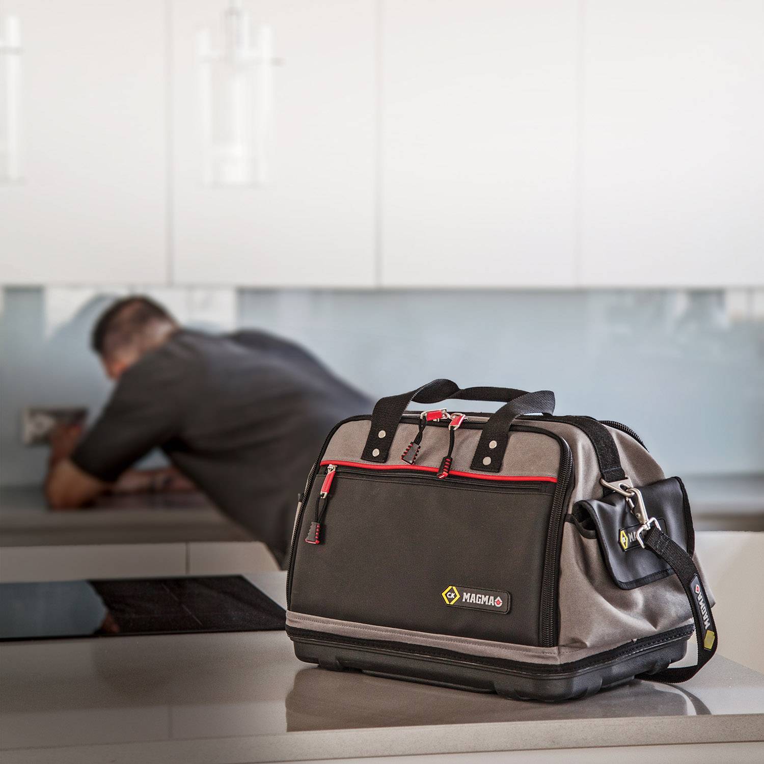 A tool bag is resting on a workbench, with a person working on a wall socket in the background.
