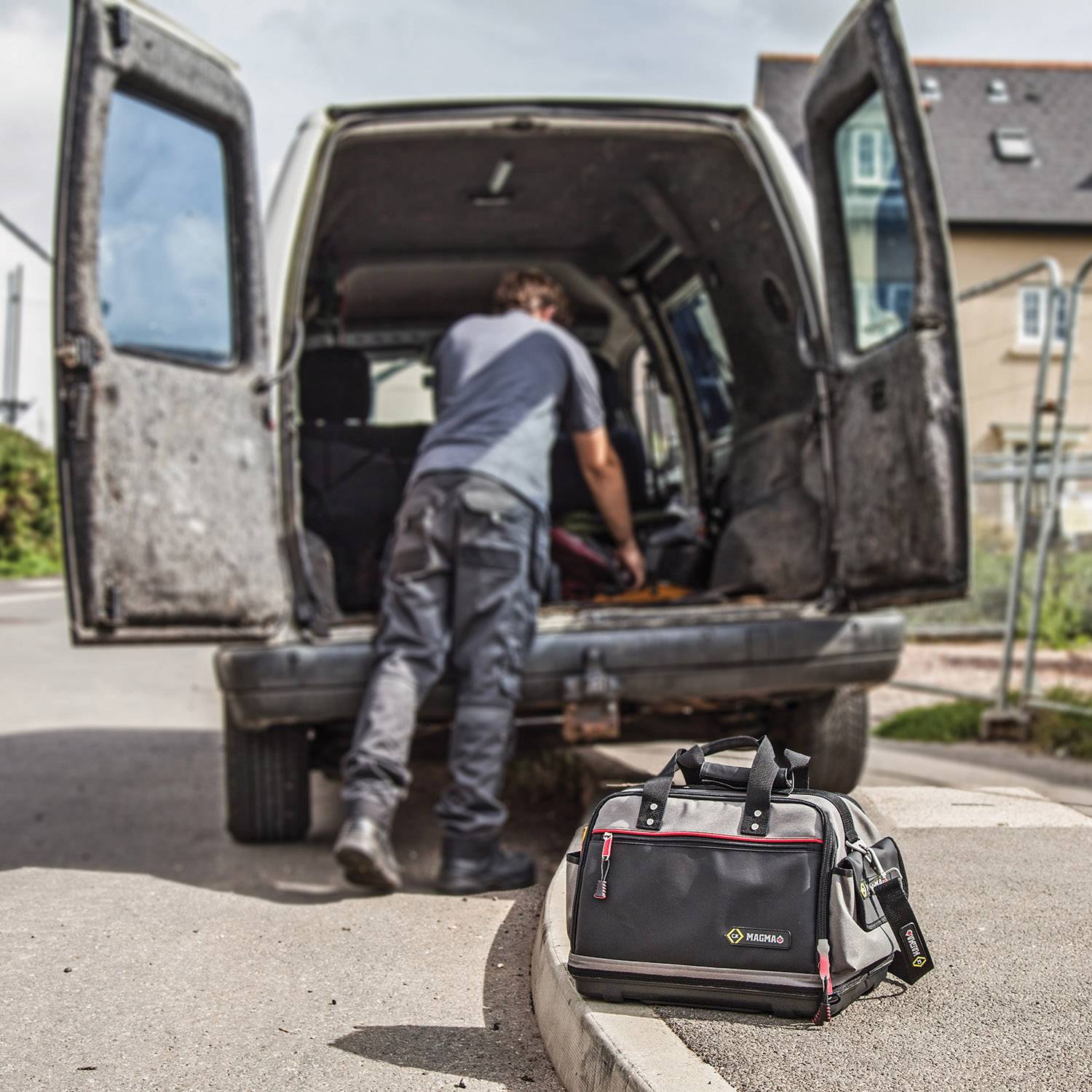A tradesman is working at the open boot of a van. A tool bag is standing on the ground in the foreground.
