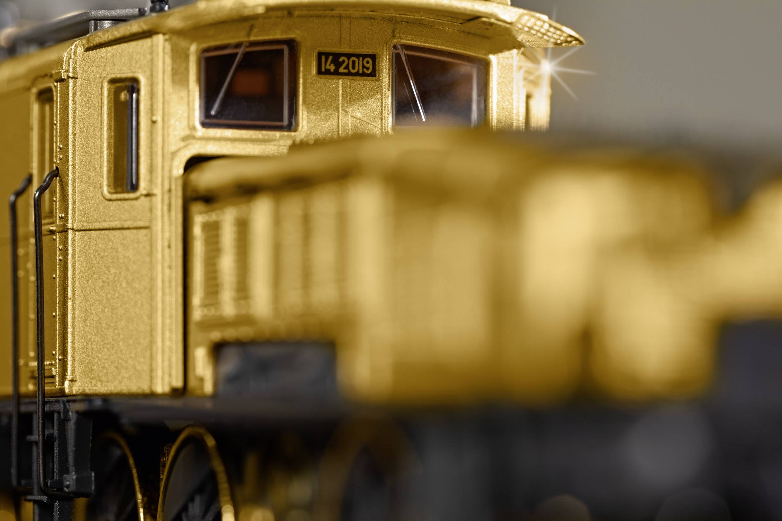 A gold-coloured train in close-up, sharp in the foreground with blurriness in the background. At the top, a number: '14 2019'.