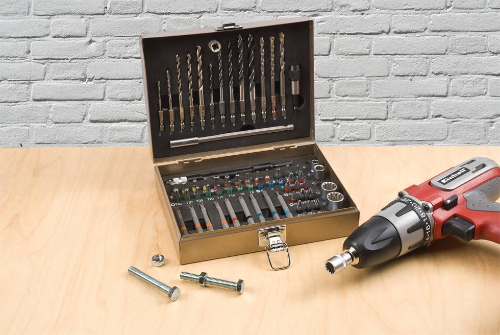 A drill set with attachments and screws is lying on a wooden table in front of a white brick wall. A red drill is placed next to the set.