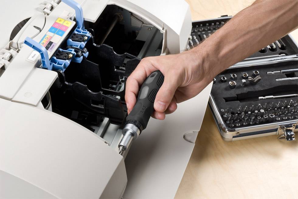 A person is repairing a printer with a screwdriver. Next to them lies a toolbox with various attachments.