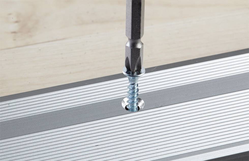 A screwdriver is driving a screw into a grooved metal rail on a wooden surface.