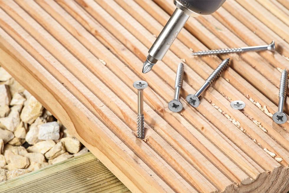A close-up of a woodworking scene with an electric screwdriver driving a screw into a wooden board resting on stones.