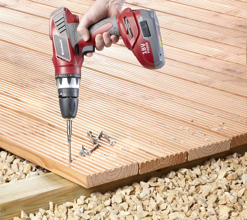 A hand holds a cordless drill and is screwing a screw into a wooden decking, surrounded by gravel stones.