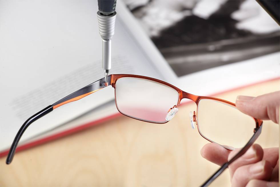 'Close-up of a hand holding a pair of glasses while a screwdriver tightens a screw on the arm. An open book is visible in the background.'