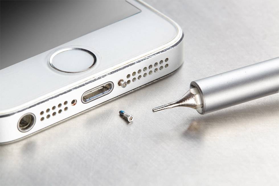 Smartphone in close-up next to a precision screwdriver on a metal surface. A small screw lies beside it.