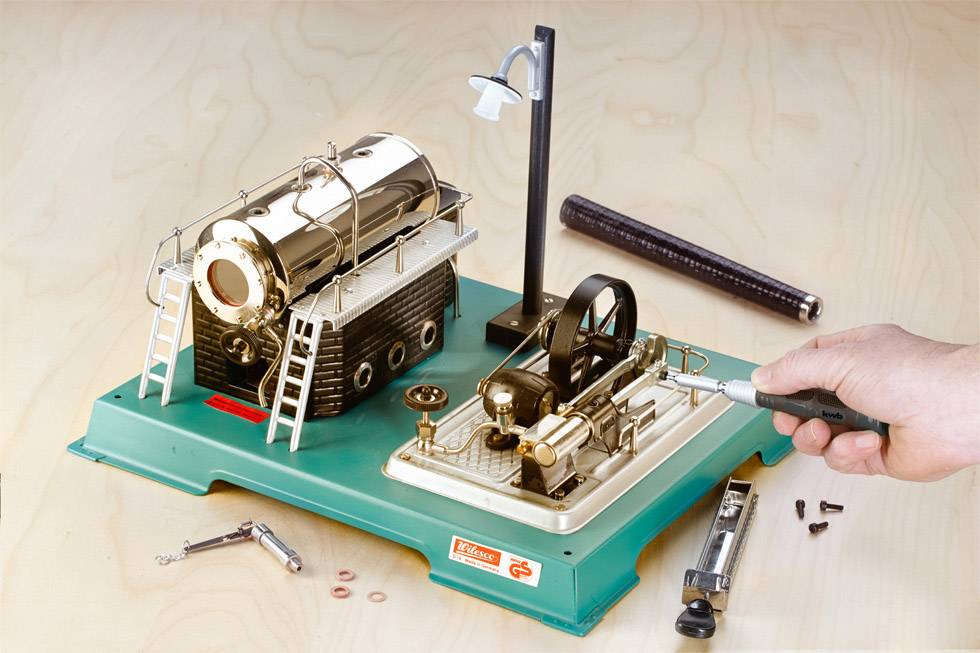 Toy steam engine set on a green base, with a gleaming boiler and a piston. Hand operating the lever to create movement.