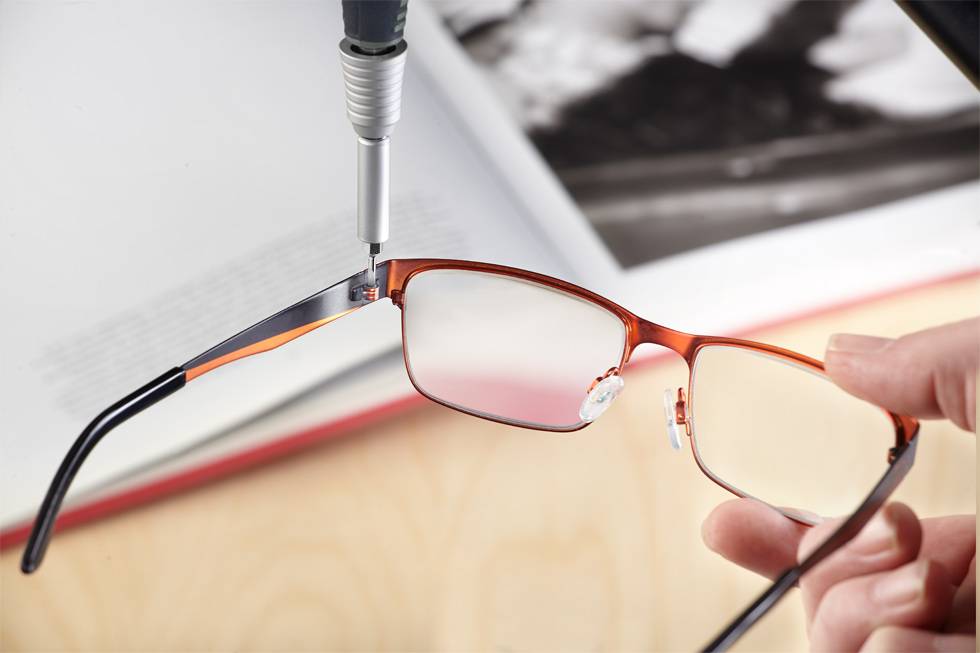 A hand is holding a pair of glasses while a screwdriver works on a frame. An open book lies on a table in the background.