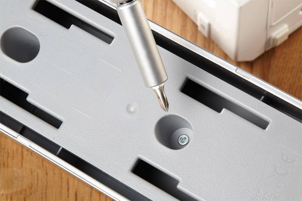 A silver screwdriver points at a screw in a white plastic cover lying on a wooden table.