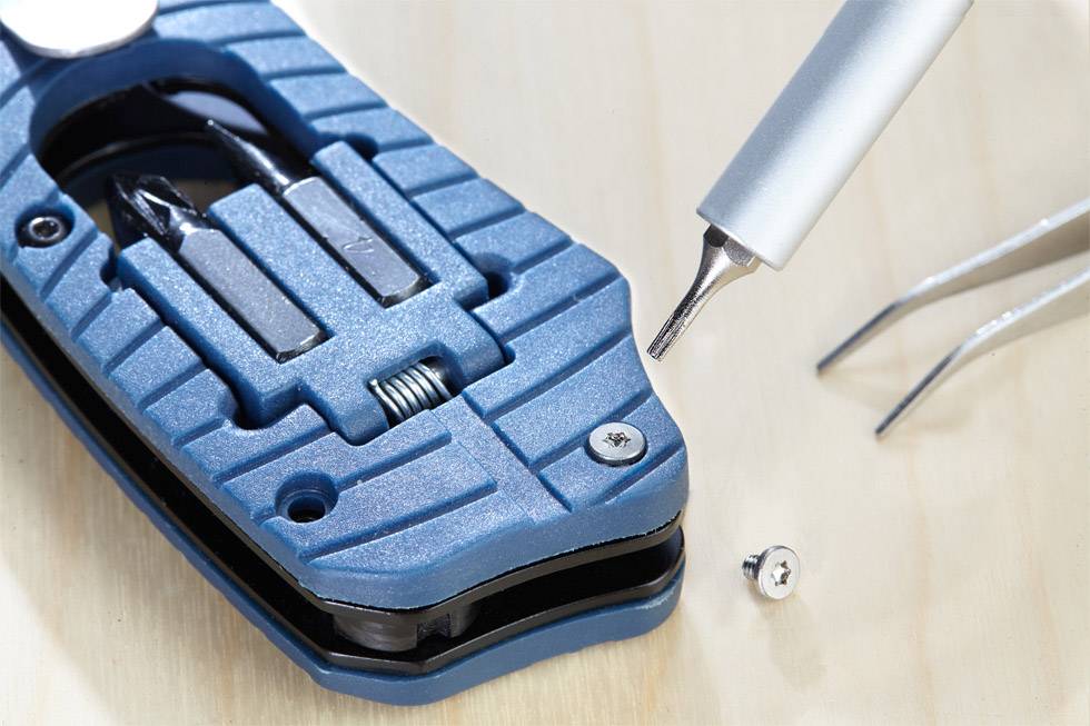 A blue screwdriver head lies open on a wooden table. A silver tip and a screw are visible beside it.