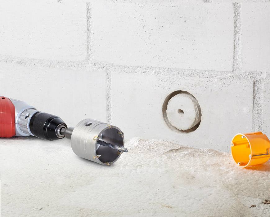 Drill with hole saw drilling a hole into a concrete wall; concrete dust and hole visible in the foreground.