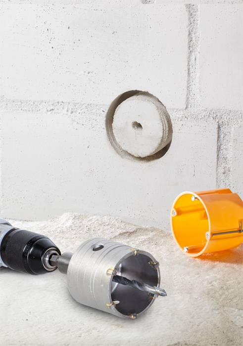 A drill with a drill bit beside it, a drilled hole in a concrete wall, and an orange drill bit on the surface.