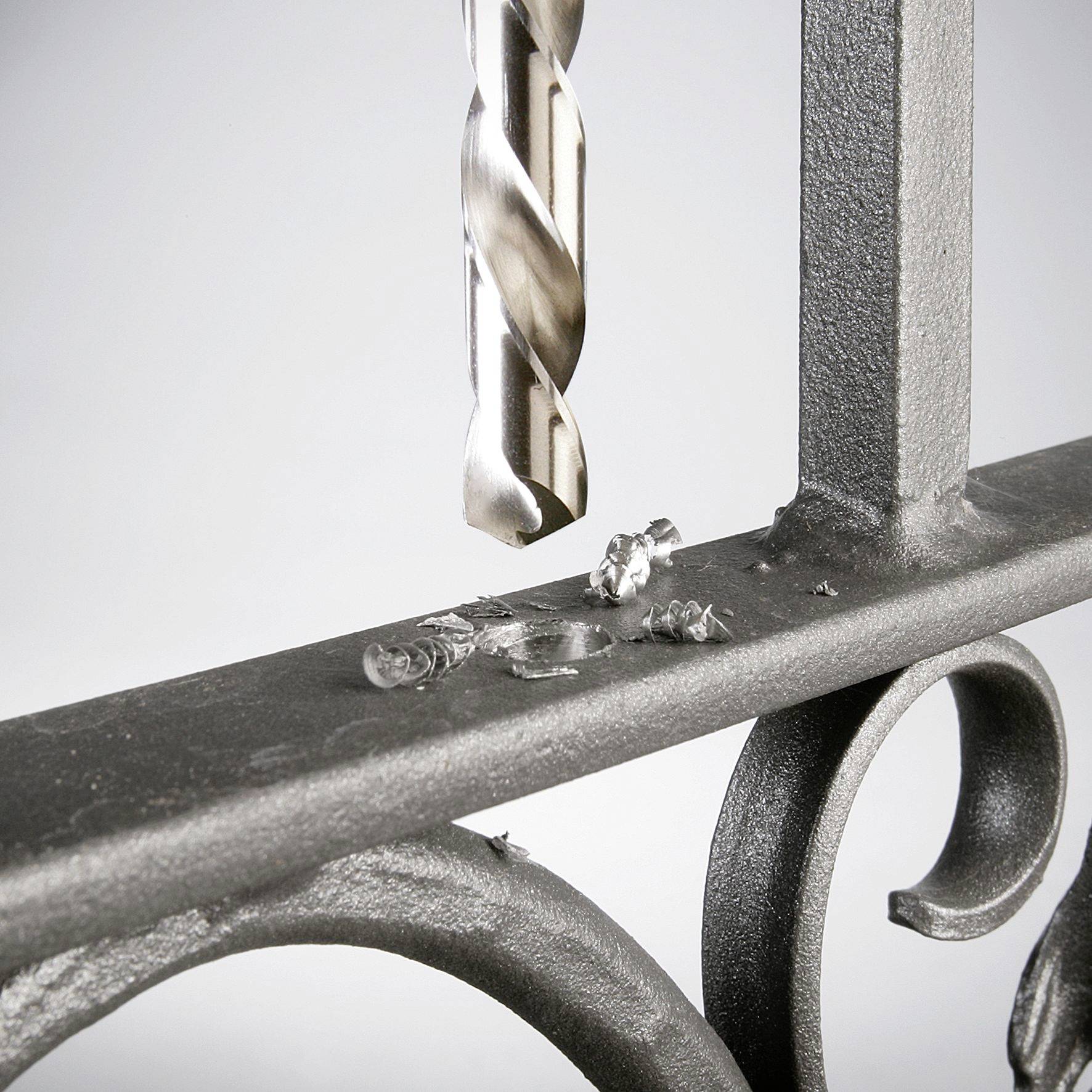 A drill penetrates a metal component that is part of a decorative handrail. Silver shavings lie at the drilling site.
