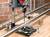 Drill positioned on a workbench, machining metal pipes, with metal shavings visible. Brick wall in the background. Tools and components positioned nearby.