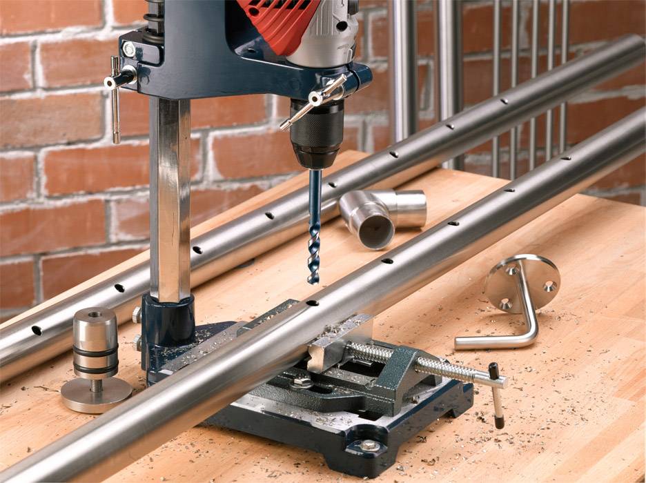 Electric drill on a wooden workbench drilling holes into a metal pipe, brick wall in the background. Metal shavings are scattered around. Tools are visible.