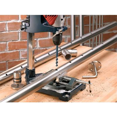 Electric drill on a wooden workbench drilling holes into a metal pipe, brick wall in the background. Metal shavings are scattered around. Tools are visible.