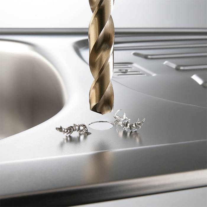 A drill attachment is drilling a hole in a metal surface beside a sink, surrounded by metal shavings.