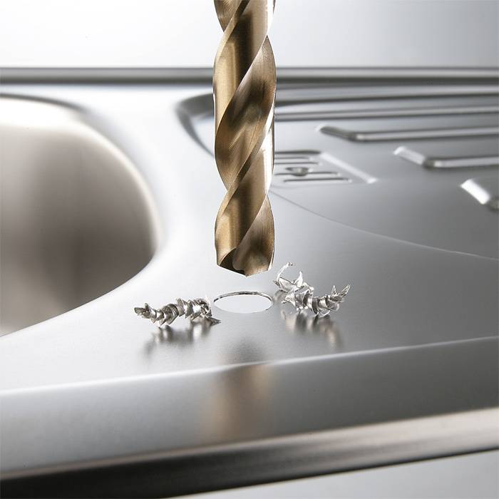 A metal drill is boring a hole into a stainless steel sink, surrounded by metal shavings.