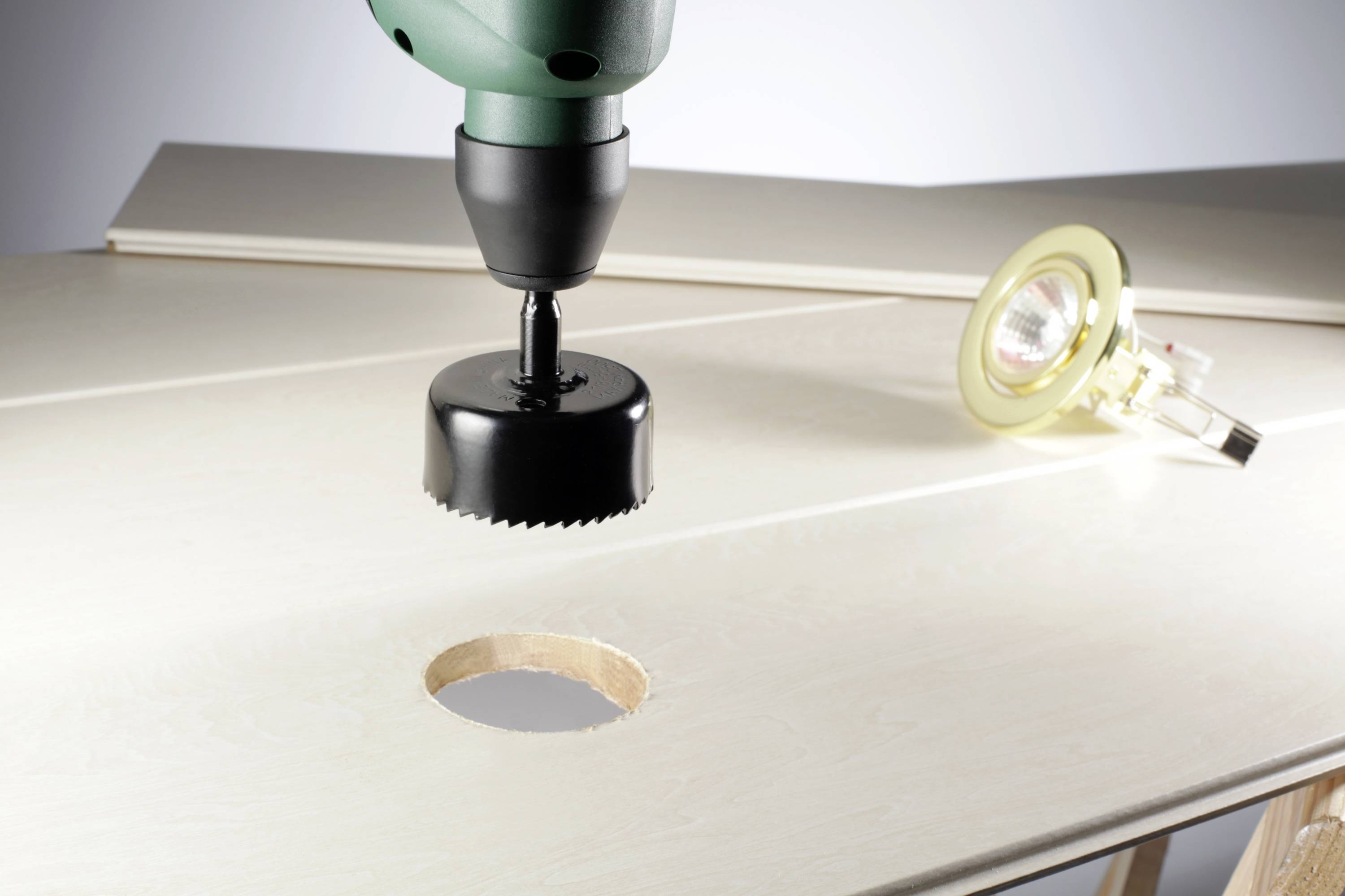 A drill with a hole saw is drilling a circular hole in a wooden board. A small lamp is lying to the right.