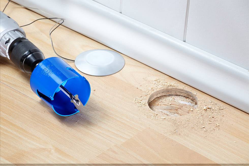 A blue drill bit next to a freshly drilled hole in a wooden floor; wood shavings are scattered around the hole.