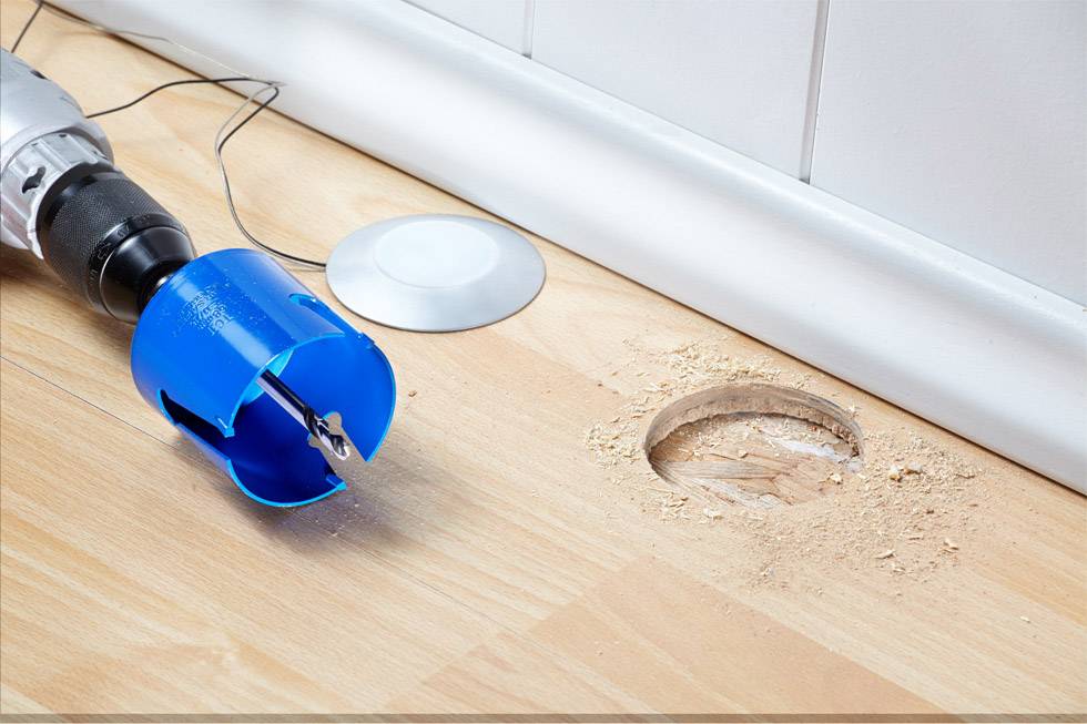 A drill with a blue drill bit lies next to a freshly drilled hole in a wooden floor. Wood shavings are scattered around the hole.
