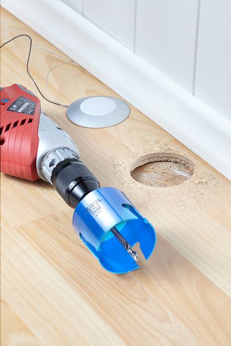 Drill with attachment pierces wooden floor, finished hole visible.