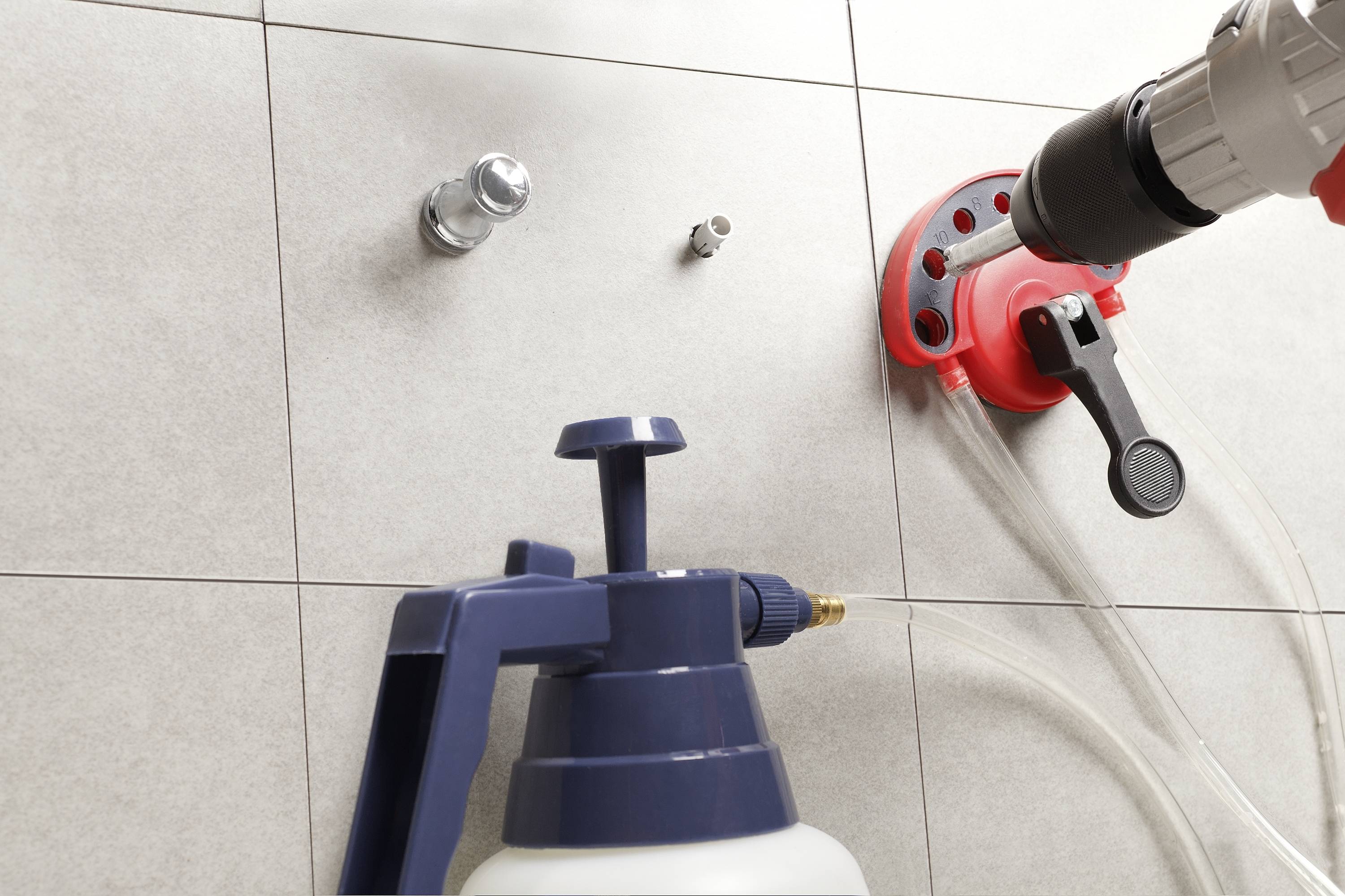 A tradesman is drilling holes in a tiled wall. A red device is securing the drill. A spray bottle stands in the foreground.