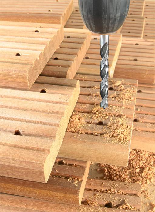 A drill is boring a hole through stacked wooden planks, with wood shavings falling all around.