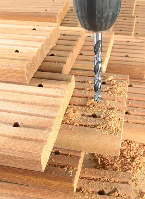 An electric drill creates holes in stacked wooden planks, with wood shavings visible.
