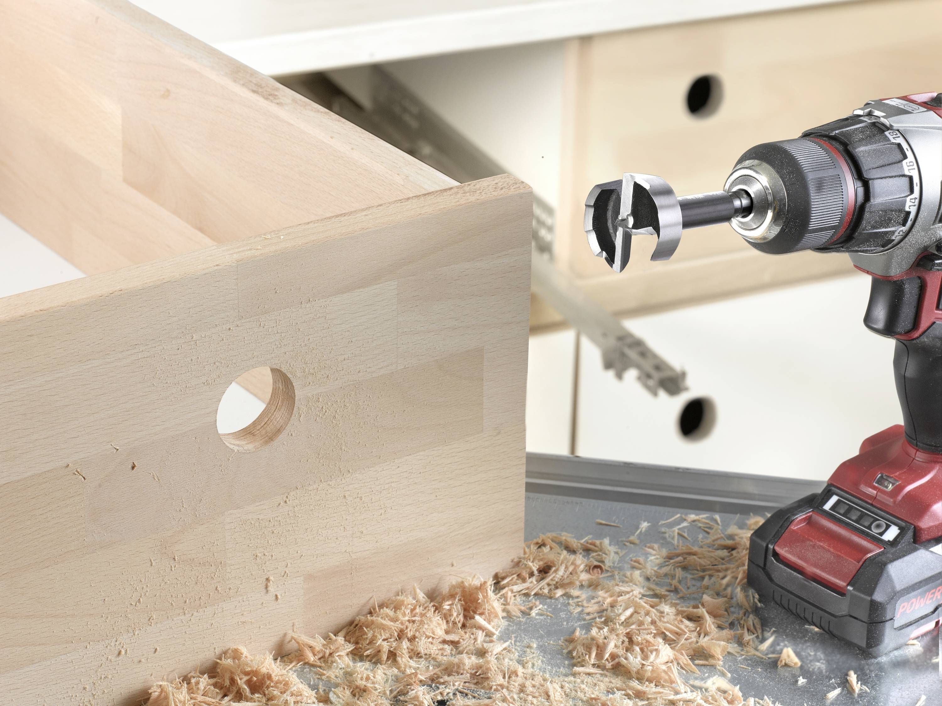A cordless drill with a Forstner bit is drilling a hole in a wooden board, with wood shavings falling to the floor. Drawers are visible in the background.