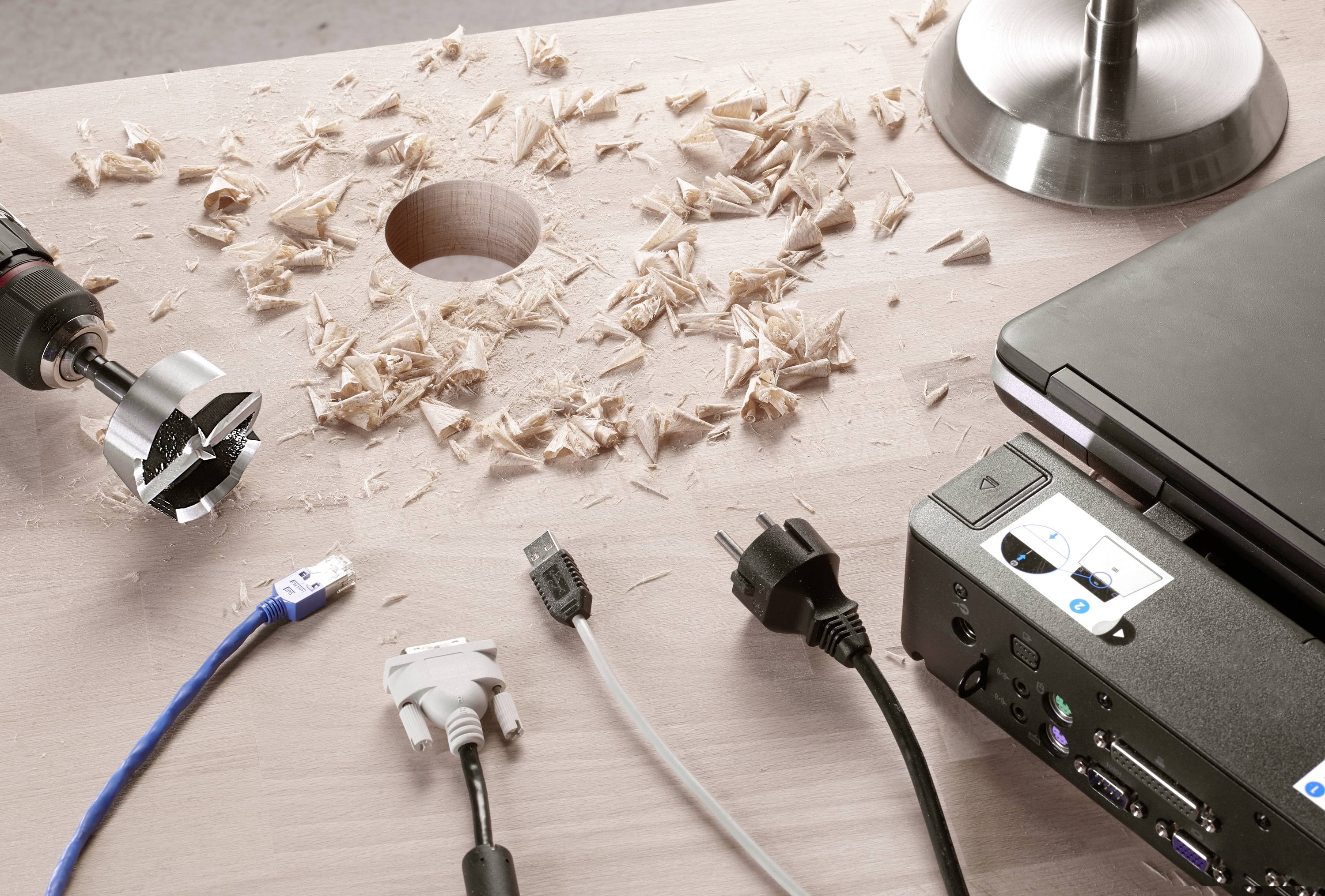 A workbench with wooden dowels and wood shavings, a drill, laptop and various cables, including power and network connections.
