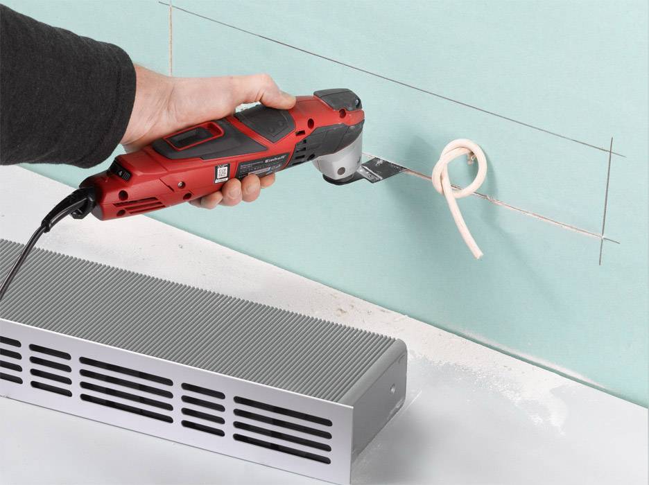 A person is cutting a rectangle into a green wall using a red multi-purpose tool, with wood shavings coming out.