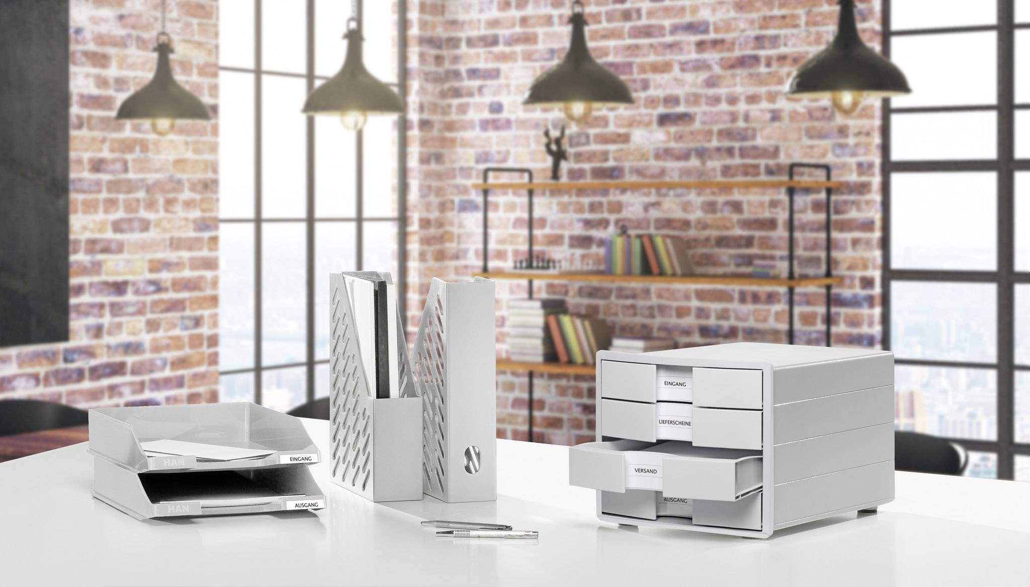 Office with modern white desk organisers, including paper trays, vertical file holders and drawer boxes in front of a brick wall.
