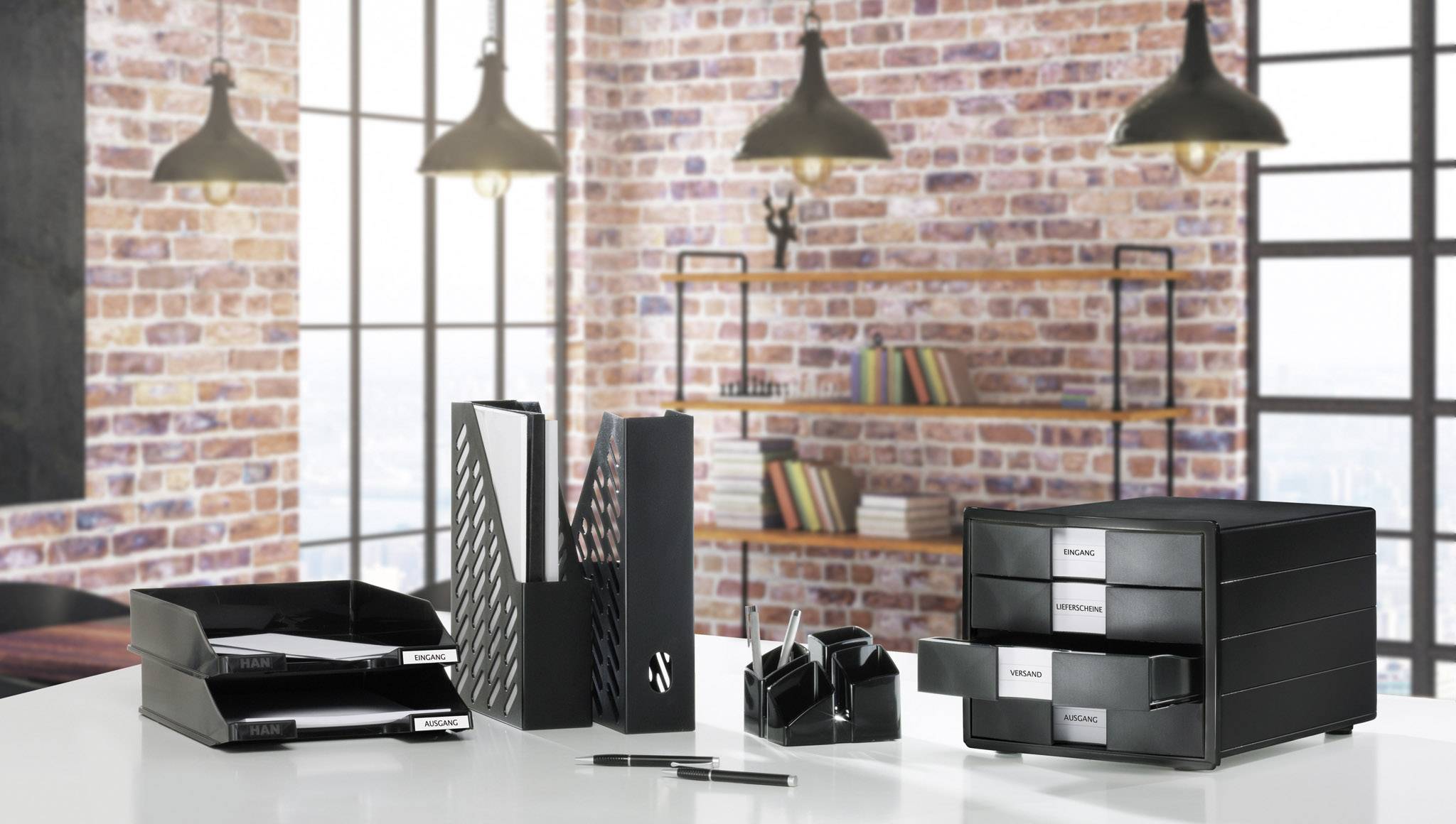 Office accessories set on a desk in front of a brick wall. Includes filing trays, folders and a pen holder in a modern design.