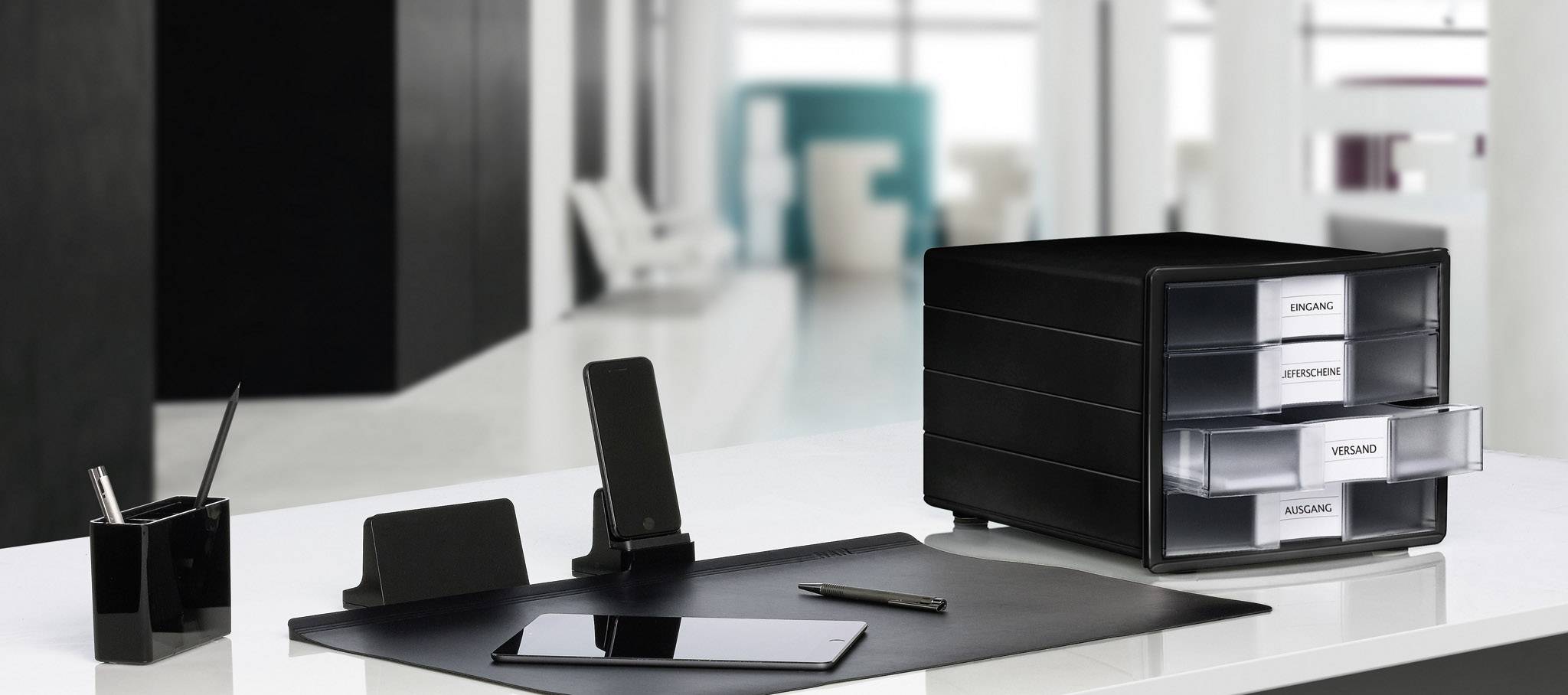 A modern desk organiser with multiple black compartments and labels, surrounded by office supplies such as a pen and tablet.