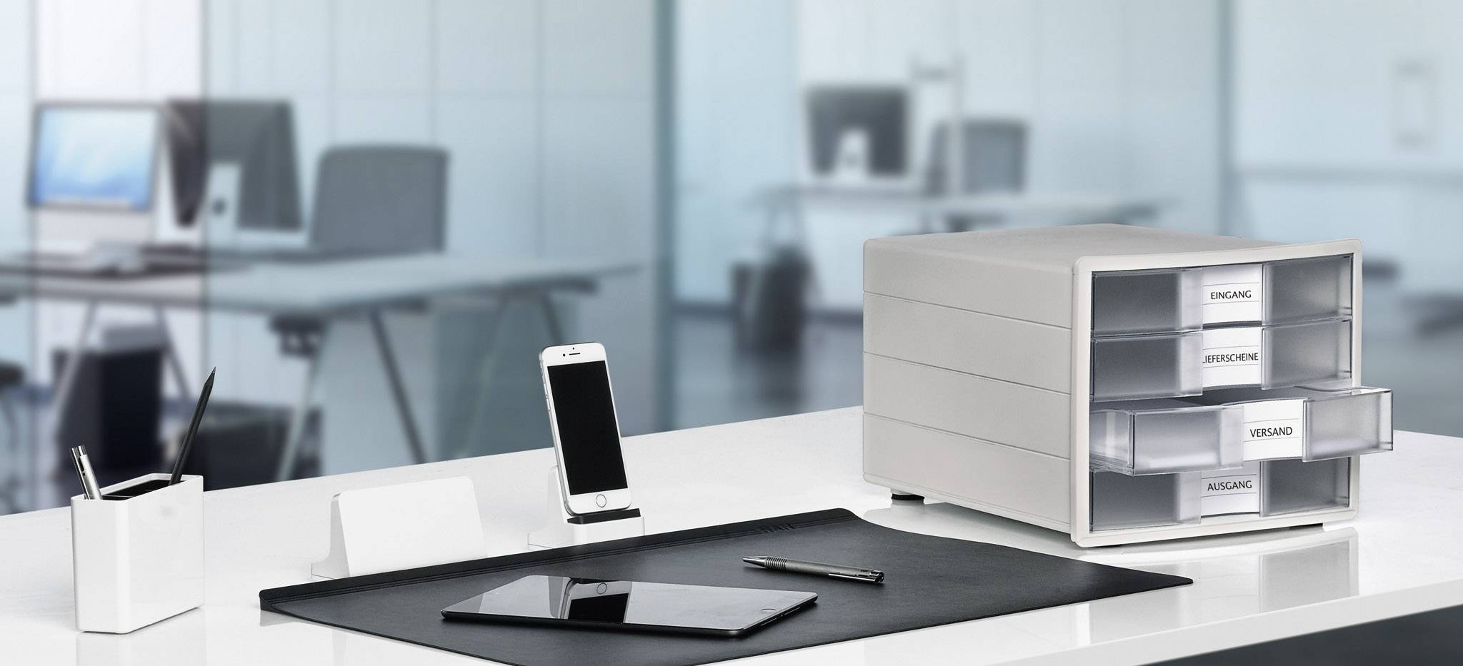 Modern desk in an office with a smartphone, tablet, pencil pot and drawer unit. Blurred workstations are visible in the background.
