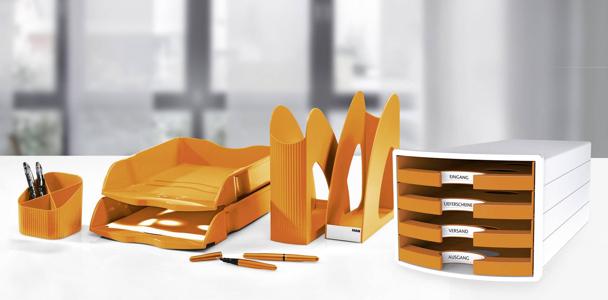 Orange desk organisers and filing trays on a table with a blurred office background. Includes pen holders, letter trays, and document stands.