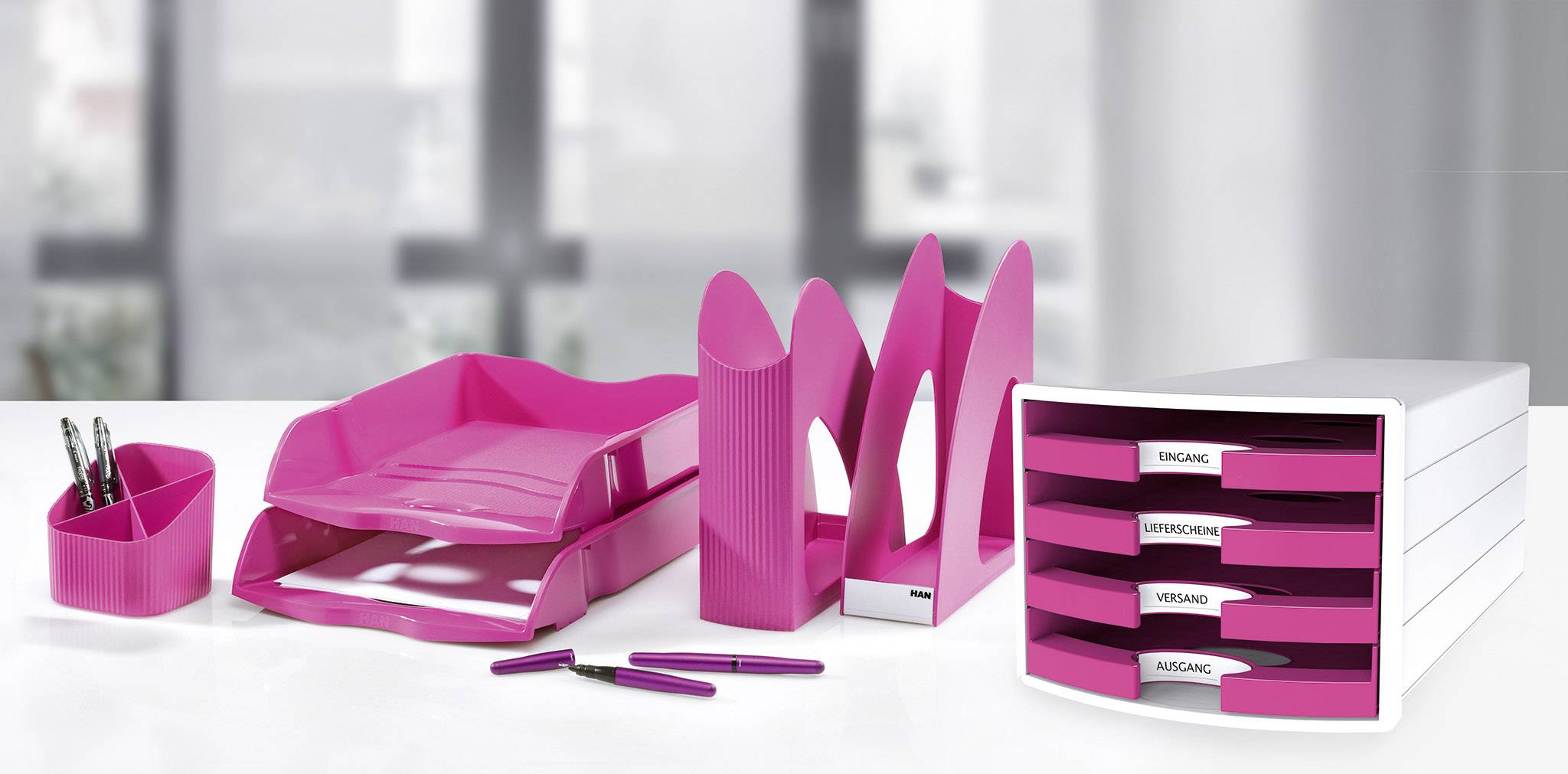 An organisational set on a desk with pink office accessories: pen holder, letter trays, folders and drawer box.