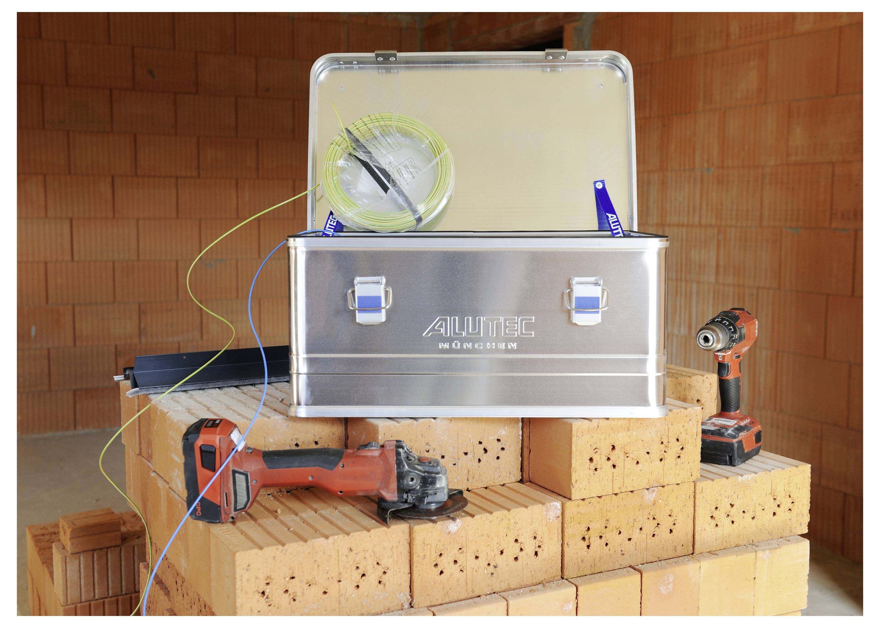 A metal storage box labeled 'Alutec' sits open on a pile of bricks, with tools like a drill and measuring tape visible around it.