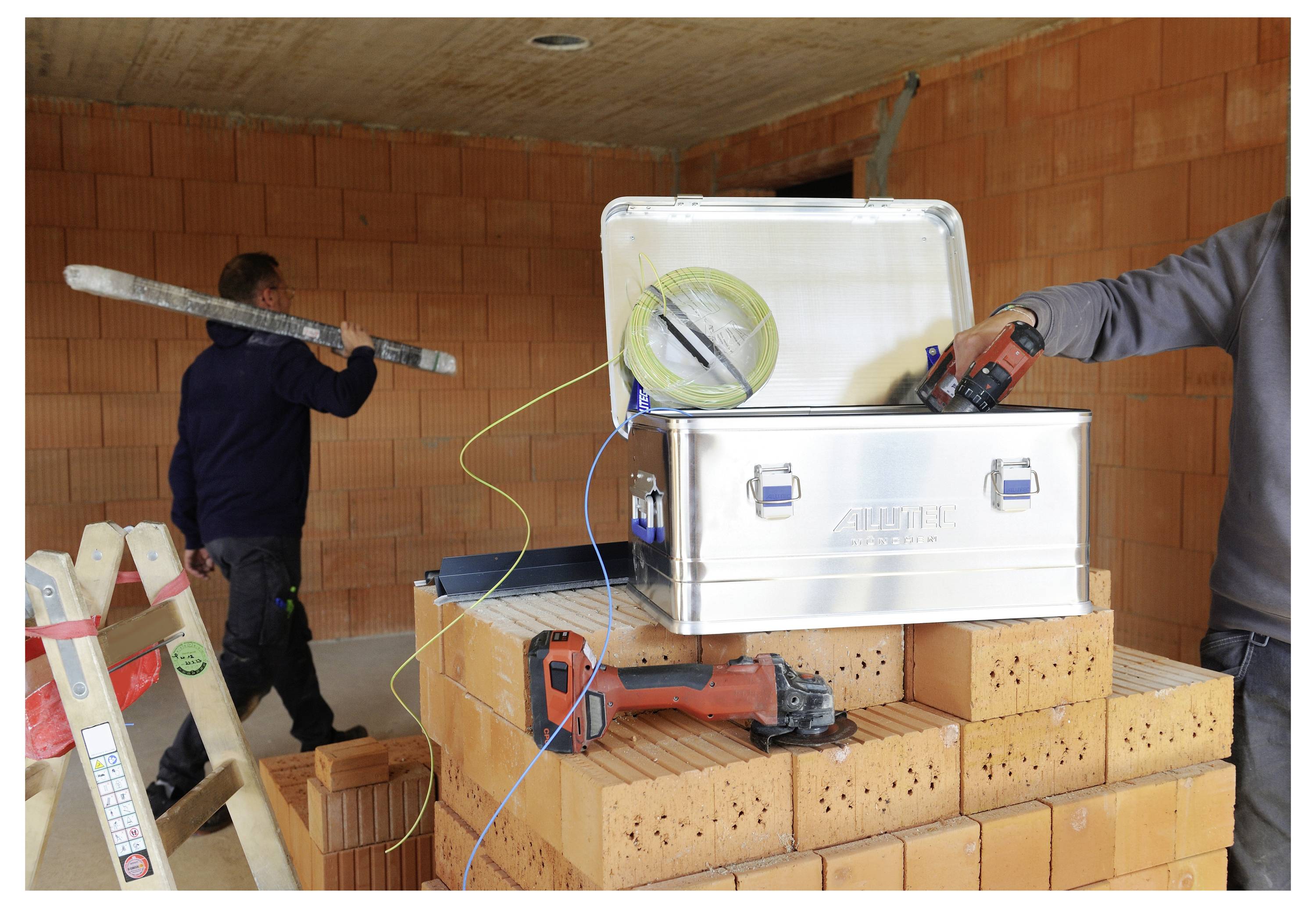 Two workers in a construction area with brick walls. One holds a tool, and a toolbox is open on a stack of bricks.