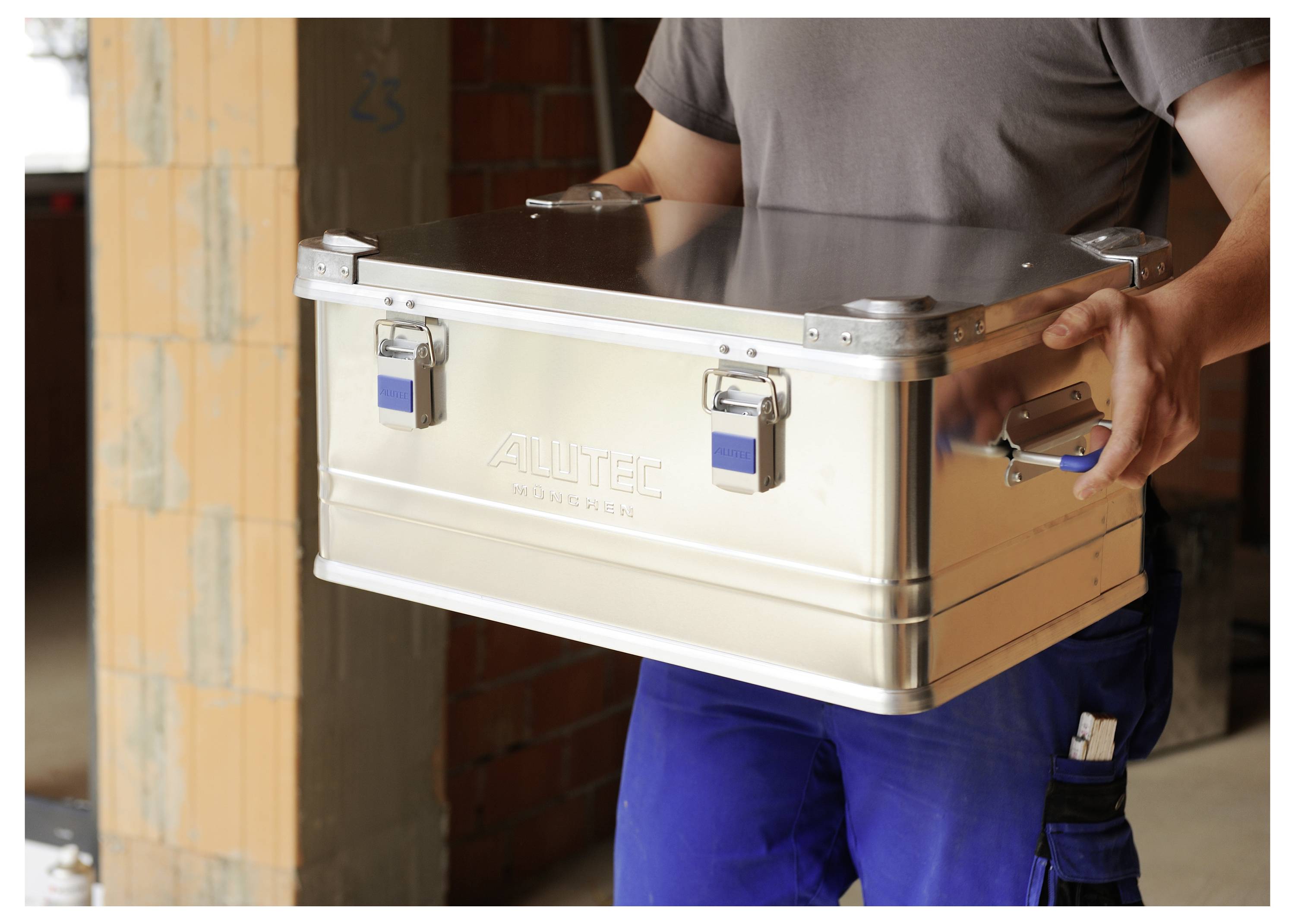 A person holding a metal storage box with side handles, standing in a partially constructed building with exposed brick walls.