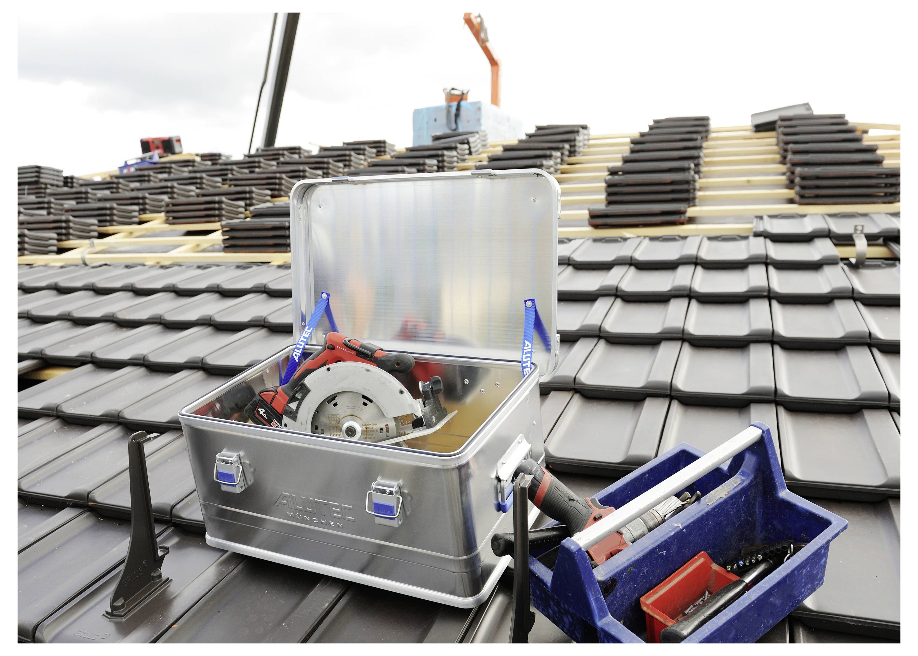 A metal toolbox with a circular saw and red handles is open on a tiled roof, with a blue container holding tools beside it.